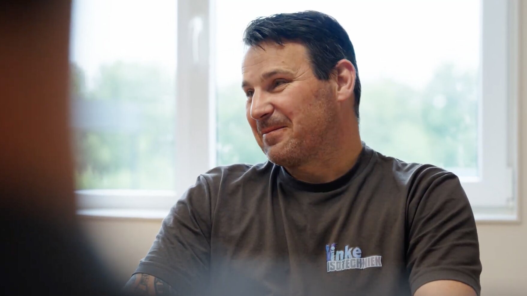 Medewerker in een T-shirt van Vinke Isotechniek glimlacht tijdens een gesprek aan tafel